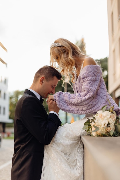 Beau Jeune Couple De Mariage En Costume Noir Et Robe Blanche Se Promener Dans La Ville S'embrasser Et Profiter Sur Fond De Rue Portrait De Mariée Heureuse Avec Bouquet Et Marié Amoureux Jour Du Mariage Vacances En Famille