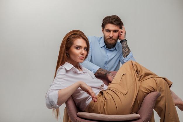 Beau jeune couple à la mode dans des vêtements à la mode dans le studio Jolie fille rousse dans une chemise blanche est assise sur une chaise à côté d'un bel homme hipster avec un tatouage et une barbe