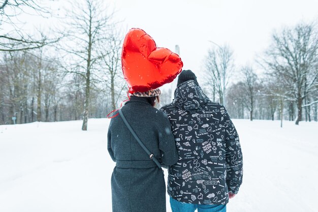 Un beau jeune couple à la mode en vêtements d'hiver avec un cœur de ballon d'air rouge marchant ensemble dans un parc enneigé