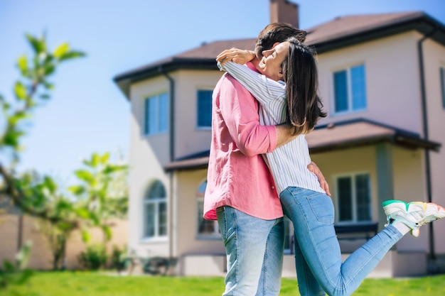 Beau jeune couple moderne et excité, amoureux, debout devant la nouvelle grande maison avec les clés en mains et s'embrassant pendant qu'ils célèbrent cet achat