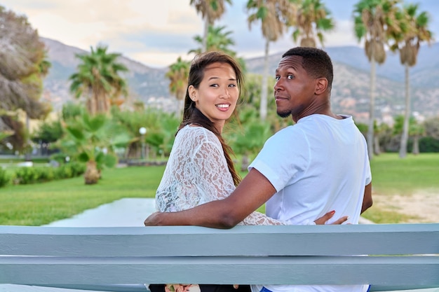 Beau jeune couple multiethnique assis ensemble sur un banc dans un parc tropical