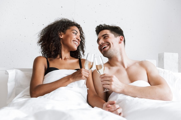 Beau jeune couple multiethnique heureux se détendre dans son lit sous une couverture, boire du champagne mousseux dans des verres