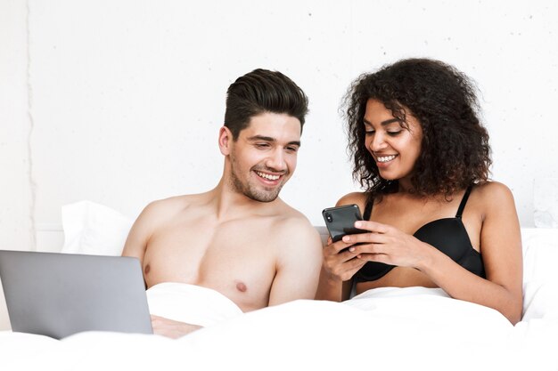 Beau jeune couple multiethnique heureux se détendre dans son lit sous une couverture, homme utilisant un ordinateur portable, femme tenant un téléphone mobile