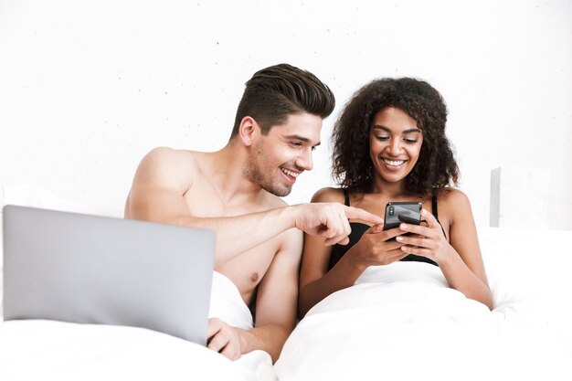 Beau jeune couple multiethnique heureux se détendre dans son lit sous une couverture, homme utilisant un ordinateur portable, femme tenant un téléphone mobile