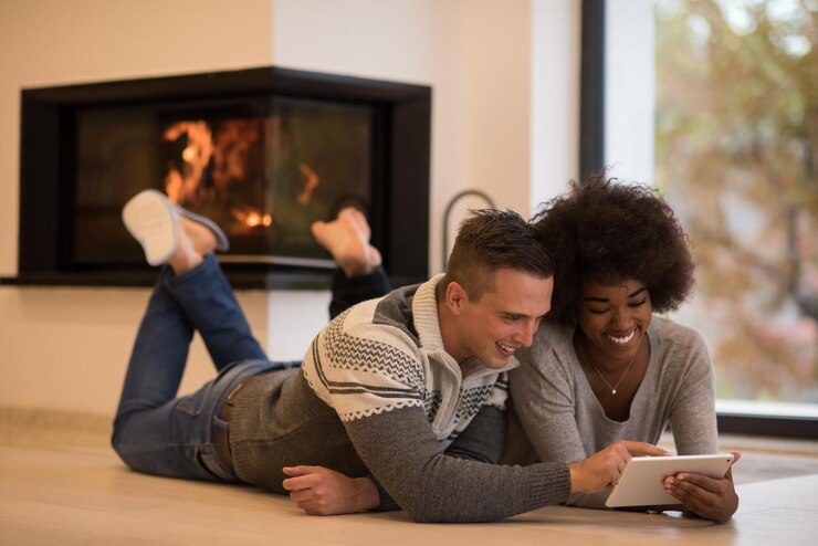Photo beau jeune couple multiethnique utilisant un ordinateur tablette sur le sol de leur maison de luxe devant la cheminée le jour de l'automne
