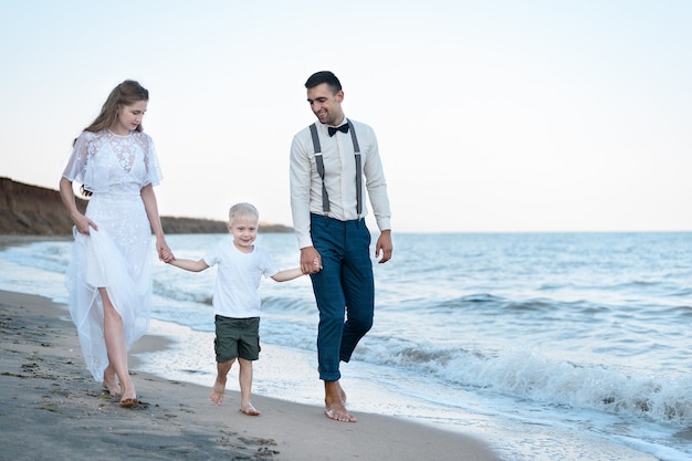 Beau jeune couple avec petit garçon se promène sur la plage. Les parents gardent les mains de leur fils.