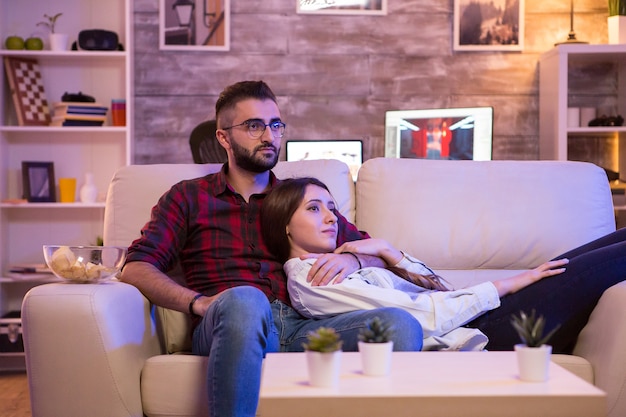 Beau jeune couple regardant leur émission de télévision préférée tout en se relaxant sur le canapé la nuit.