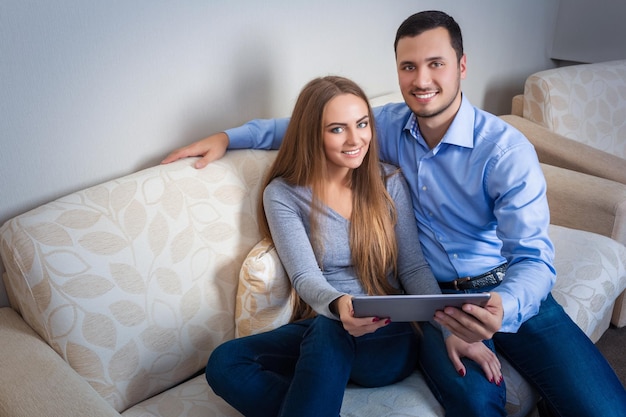 Beau jeune couple riant, assis sur un canapé, avec tablette électronique et regardant la caméra.