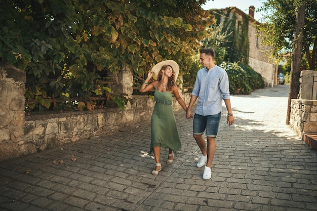 Un beau jeune couple s'amuse et danse dans la rue en marchant le long d'une ville méditerranéenne. Ils sont appréciés en journée ensoleillée d'été, se tenant la main et souriant.