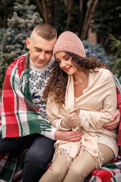 Beau jeune couple souriant heureux enveloppé dans une couverture écossaise Arbres de Noël verts sur le fond Les vacances d'hiver En attendant la nouvelle année