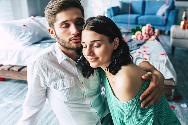 Beau jeune couple tendre et charmant s'embrassent et s'étreignent assis sur le sol de la chambre avec un fond romantique. La saint Valentin. Anniversaire. Date. Notion d'amour