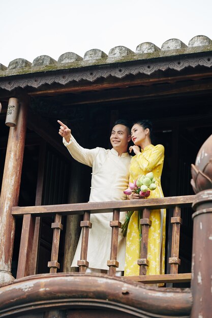 Beau Jeune Couple Vietnamien En Robes Traditionnelles Ao Dai Debout Sur Le Balcon D'un Bâtiment En Bois Et Profitant De La Vue