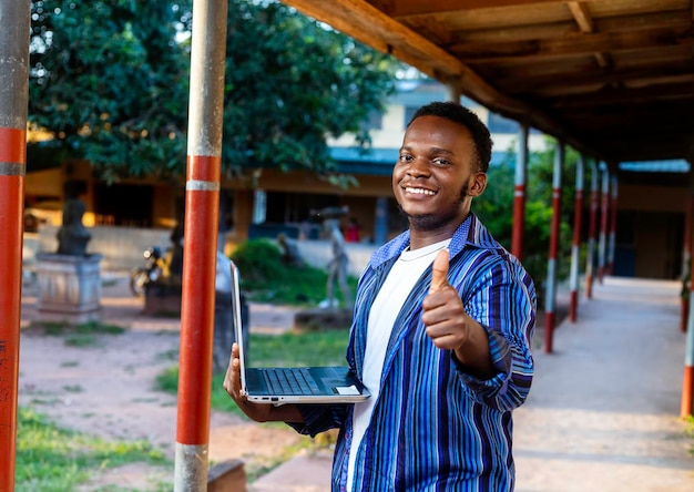 Beau jeune écolier afro-américain tenant un ordinateur portable tout en donnant les pouces vers le haut