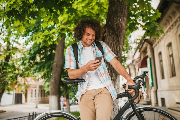 Beau jeune étudiant heureux aux cheveux bouclés portant une chemise avec sac à dos assis sur le vélo dans la rue en naviguant sur les réseaux sociaux sur téléphone mobile ayant un regard joyeux se préparant pour l'université