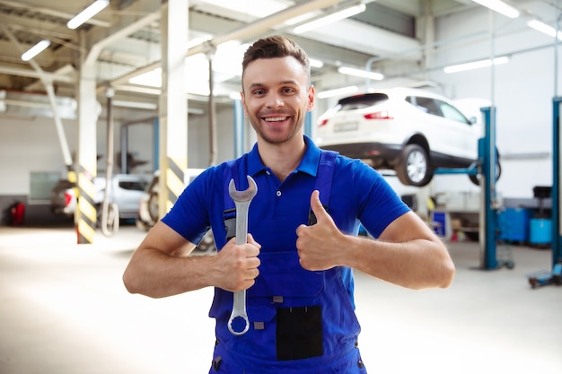 Beau jeune et expérimenté réparateur automobile confiant en salopette de travail posant dans le contexte de voitures levées dans un service de voiture