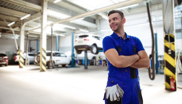 Beau jeune et expérimenté réparateur automobile confiant en salopette de travail posant dans le contexte de voitures levées dans un service de voiture