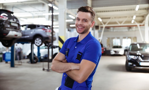 Beau jeune et expérimenté réparateur automobile confiant en salopette de travail posant dans le contexte de voitures levées dans un service de voiture