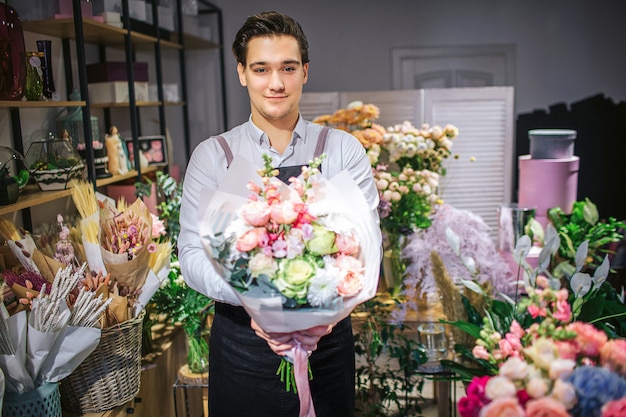 Beau jeune fleuriste mâle se tient et regarde. Il tient un bouquet de fleurs dans les mains. Guy se tient à l'intérieur. Il est gai.