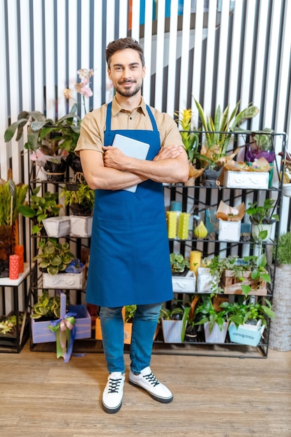 Beau jeune fleuriste tenant une tablette numérique et souriant à la caméra tout en travaillant dans un magasin de fleurs