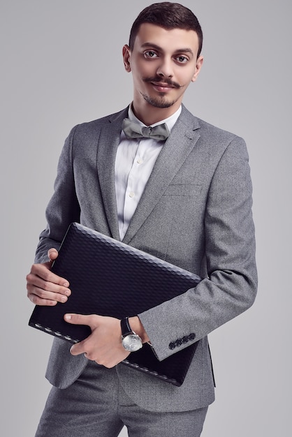 Beau jeune homme d'affaires arabe avec moustache en costume gris tient un ordinateur portable