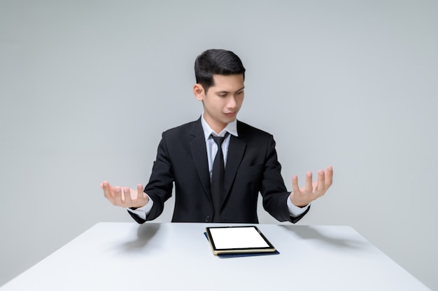 Photo beau jeune homme d'affaires assis à une table et levant les deux mains et en regardant ses mains. bel homme avec tablette et table isolé sur blanc