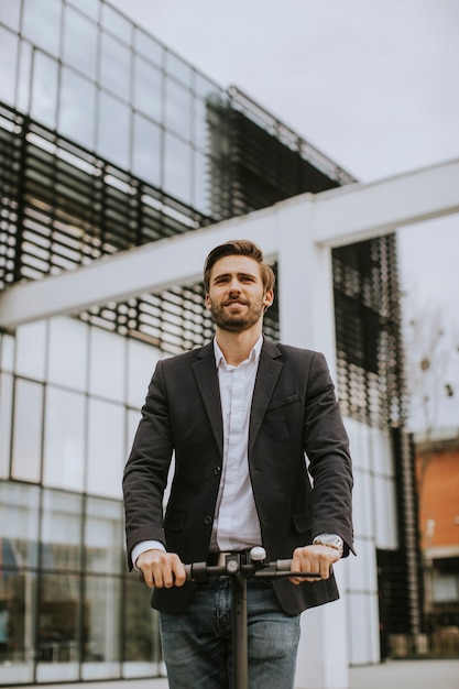 Beau jeune homme d'affaires dans un vêtement décontracté équitation un scooter électrique par un immeuble de bureaux lors d'une réunion d'affaires