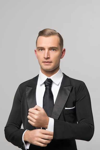 Beau jeune homme d'affaires ou gentleman aux cheveux à la mode en costume formel noir de veste, chemise et cravate sur fond de studio gris