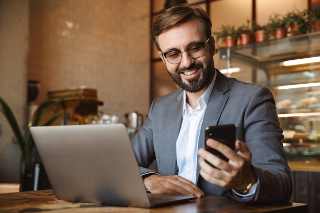 Beau jeune homme d'affaires habillé en costume travaillant sur un ordinateur portable, assis au café à l'intérieur, tenant un téléphone mobile