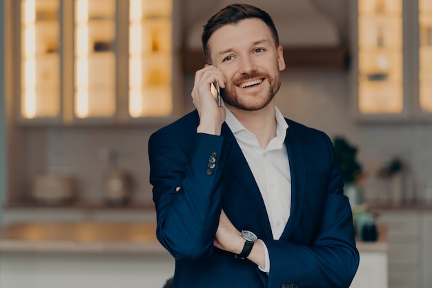 Beau jeune homme d'affaires heureux en costume classique parlant au téléphone portable et souriant, ayant une conversation agréable tout en se tenant dans la chambre de son appartement. Concept d'appel d'affaires