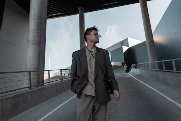 Beau jeune homme d'affaires avec des lunettes de soleil dans un costume gris à la mode avec un sac à main se promène dans la ville près d'un immeuble moderne