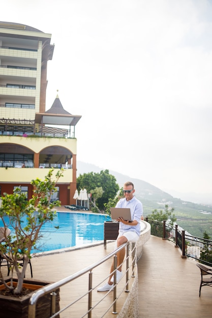 Beau jeune homme d'affaires réussi travaillant avec un ordinateur portable regarde la mer Méditerranée. Il porte une chemise et un short blanc. Travail à distance en vacances. Concept de vacances