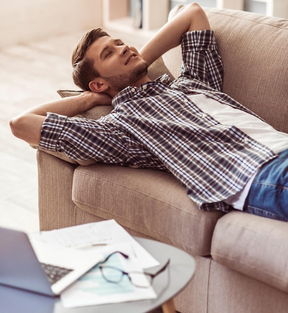 Beau jeune homme d'affaires se repose sur un canapé tout en travaillant avec un ordinateur portable à la maison