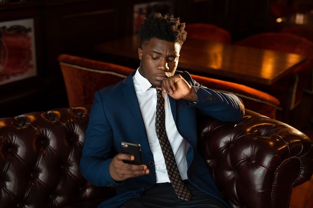 Beau jeune homme africain en costume à l'intérieur avec téléphone mobile