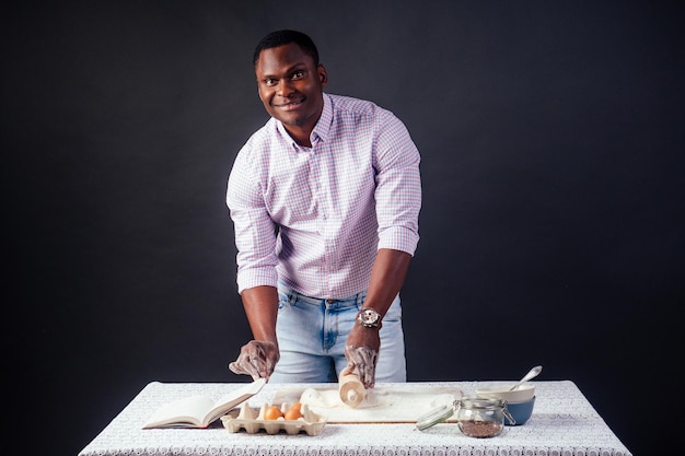 Beau et jeune homme afro-africain préparant des gâteaux faits maison Tarte américaine à partir de mains de pâte fraîche sales par la farine, sur la table se trouvent des œufs, un rouleau à pâtisserie et un livre de recettes sur fond noir dans le studio