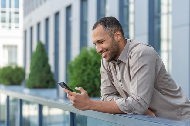 Un beau jeune homme afro-américain heureux se tient sur la véranda d'un immeuble tenant un téléphone