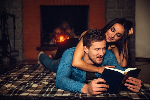 Beau jeune homme allongé sur le sol et lire le livre ouvert. Sa copine est au-dessus de lui aussi. Ils passent du temps près de la cheminée.