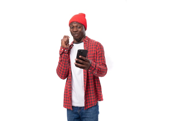 Beau jeune homme américain en couvre-chef rouge assis sur internet à l'aide d'un téléphone intelligent sur blanc isolé