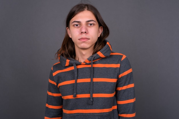 Beau jeune homme androgyne aux cheveux longs portant un sweat à capuche contre le mur gris