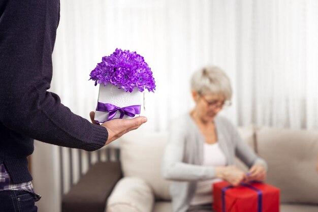 Photo un beau jeune homme apporte de belles fleurs violettes à sa mère qui tient dans ses bras un autre cadeau.
