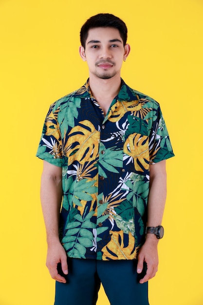 Beau jeune homme asiatique en chemise hawaïenne colorée debout à la recherche d'appareil photo. Portrait en studio de la moitié du corps sur fond jaune. Des gens détendus et heureux dans le concept d'été