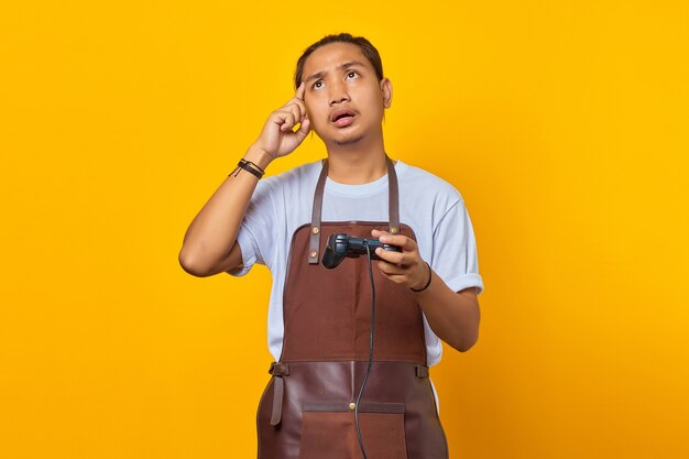 beau jeune homme asiatique portant un tablier tenant un contrôleur de jeu et pensant à quelque chose