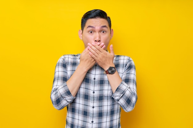 Beau jeune homme asiatique surpris en chemise à carreaux couvre sa bouche avec sa main isolé sur fond jaune concept de mode de vie des gens