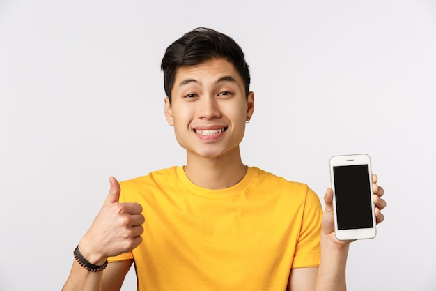 Beau jeune homme asiatique en t-shirt jaune, abandonnant le pouce et tenant le smartphone