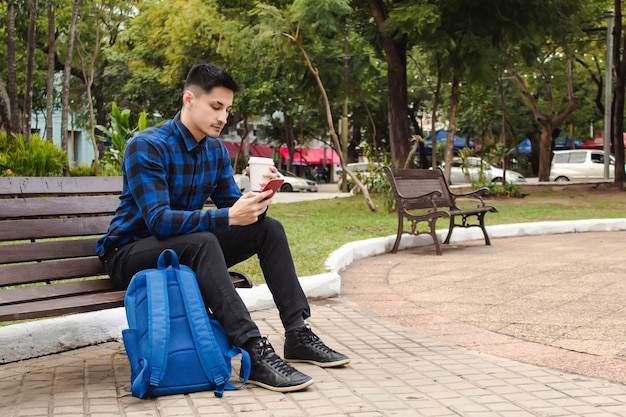 Un beau jeune homme assis sur un banc sur une place, utilisant son téléphone intelligent tout en tenant une tasse de café en papier, avec espace de copie.