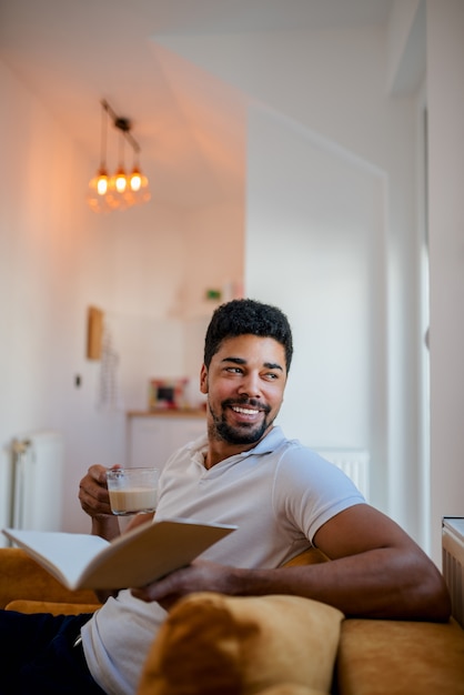 Beau jeune homme assis sur un canapé, lisant un livre et buvant du café.