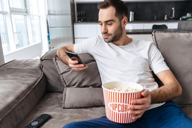 Beau jeune homme assis sur un canapé à la maison, mangeant du pop-corn, regardant la télévision, utilisant un téléphone portable