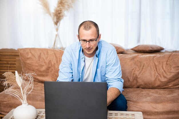 Beau jeune homme assis sur le canapé et travaillant sur ordinateur portable