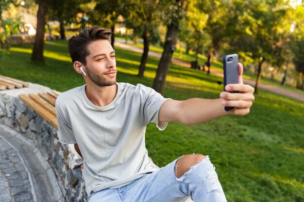 Beau jeune homme assis dans le parc à l'extérieur en prenant un selfie par téléphone portable et en écoutant de la musique avec des écouteurs