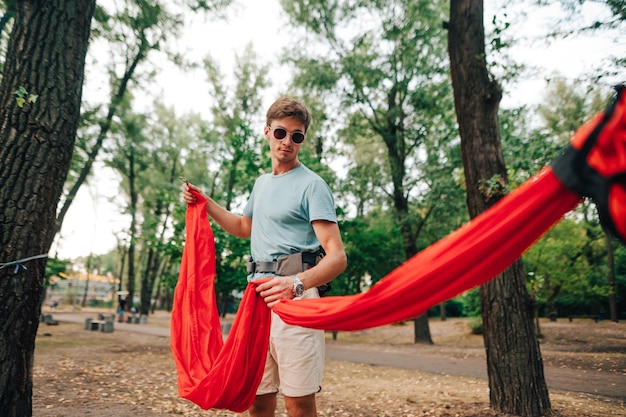 Beau jeune homme attache un hamac rouge à un arbre dans le parc pour se détendre