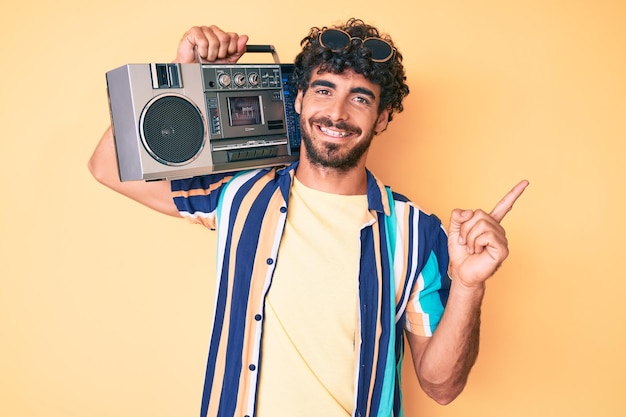 Beau jeune homme aux cheveux bouclés et ours tenant une boombox, écoutant de la musique portant un look d'été souriant heureux pointant avec la main et le doigt sur le côté